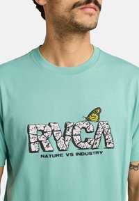 Camiseta de algodón turquesa con un gran logo de "RVCA" en negro desgastado y una mariposa amarilla, acompañado del texto "NATURE VS INDUSTRY" debajo.