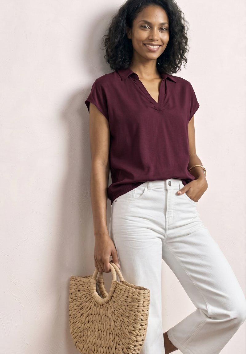 Femme aux cheveux bouclés portant un haut bordeaux et un pantalon blanc, tenant un sac à main en paille tressée, debout contre un mur clair.