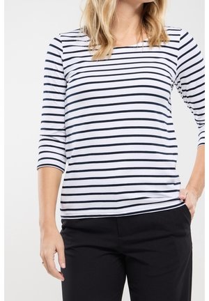 Nainen pukeutuneena valkoiseen pitkähihainen paita, jossa mustat vaakaraidat, ja mustiin housuihin, toinen käsi taskussa, yksiväristä taustaa vasten.