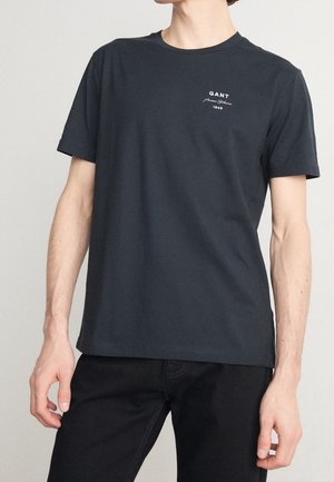 Homme portant un t-shirt GANT bleu marine foncé à manches courtes avec un logo texte sur le côté gauche de la poitrine, associé à un pantalon noir sur un fond uni.