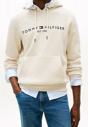 Beiger Tommy Hilfiger Hoodie mit Logo und "EST. 1985" über einem hellblau gestreiften Hemd, kombiniert mit blauen Jeans, Hände in den Taschen.