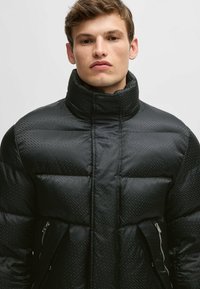 Svart vadderad jacka med ett kviltat mönster, hög krage och dragkedjefickor på sidan. Materialet har en slät, glansig yta.