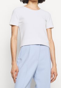 T-shirt bianca a maniche corte realizzata in tessuto liscio, con scollatura rotonda e un design leggermente cropped. Abbinata a pantaloni blu chiaro.