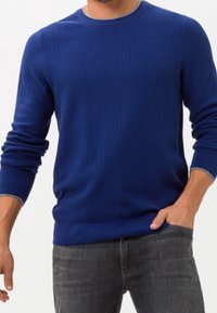 Ribbad blå stickad tröja med rund halsringning, långa ärmar och mudd. Bärs över grå jeans, vilket visar upp en figurnära design och texturerad tyg.