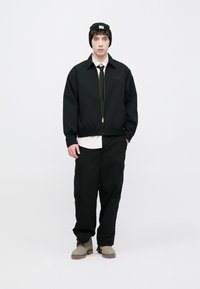 Μαύρο μπουφάν με φερμουάρ και άνετη εφαρμογή, φορεμένο πάνω από λευκό πουκάμισο, συνδυασμένο με μαύρο παντελόνι cargo και μπεζ μποτάκια. Μαύρο σκουφί με λογότυπο.