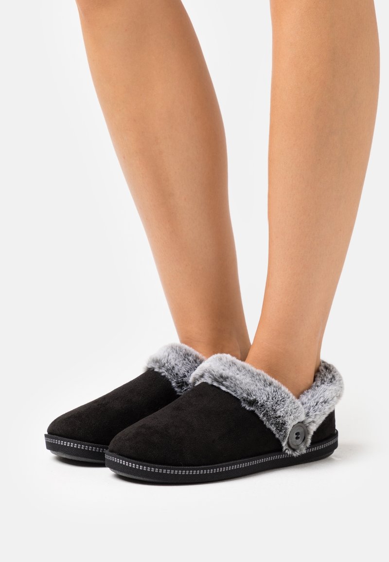 Pieds portant des chaussons noirs avec une doublure en fausse fourrure grise et un bouton décoratif, présentés sur un fond blanc uni.