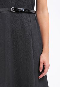 Gros plan d'une robe noire texturée avec une ceinture noire brillante et un bras droit visible contre un fond clair uni.