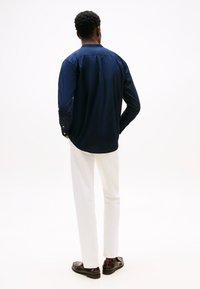 Camisa de manga larga azul marino con dobladillo curvado y puños con botones, combinada con pantalones blancos y zapatos oscuros, vista desde atrás.