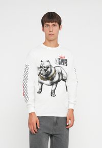 Wit overhemd met lange mouwen, voorzien van een grafisch ontwerp van een gespierde hond met een gouden ketting, zwarte details en een maaspatroon op de mouwen.