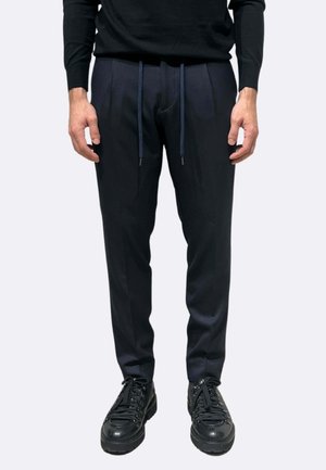Pantaloni blu navy con taglio sartoriale, caratterizzati da pieghe frontali e vita elasticizzata. Abbinati a scarpe nere stringate.