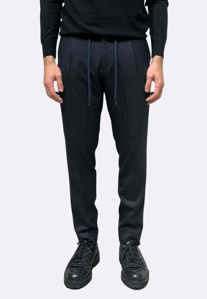 Pantaloni blu navy con taglio sartoriale, caratterizzati da pieghe frontali e vita elasticizzata. Abbinati a scarpe nere stringate.