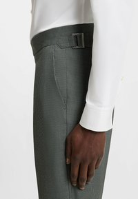 Grau karierte Hose aus strukturiertem Stoff mit einem Gürtelschlaufen und einer silbernen Schnalle. Ein weißer Hemdenbund ist oben sichtbar.