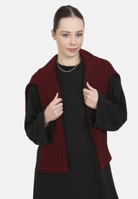 Burgundy ribbad stickad väst med öppen framsida, med en bred krage, bärs över en svart långärmad tröja. Texturerat material.