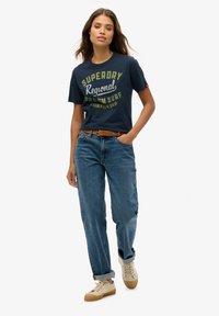 Camiseta de manga corta en navy con gráfico de "Superdry Regional Surf Championship"; combinada con vaqueros anchos de color azul claro y zapatillas beige.