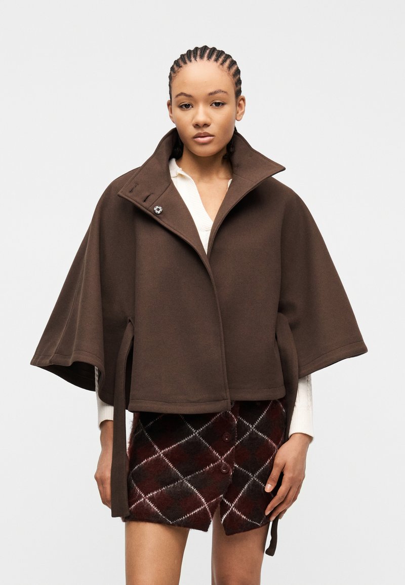 Νέα γυναίκα με πλεξούδες, που φοράει καφέ κοντό κάπα παλτό πάνω από ένα λευκό πουλόβερ και μια μπορντό καρό φούστα με κουμπιά.