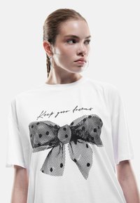 Biała koszulka z krótkim rękawem z dużą grafiką czarnej siateczkowej kokardy w kropki oraz napisem "Zachowaj swoje marzenia" w kursywie.