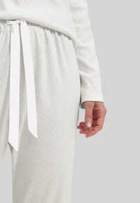 Pantalones de pijama blancos, texturizados, con un patrón de zigzag y un cordón de cinta blanca en la cintura. La tela parece suave y ligera.