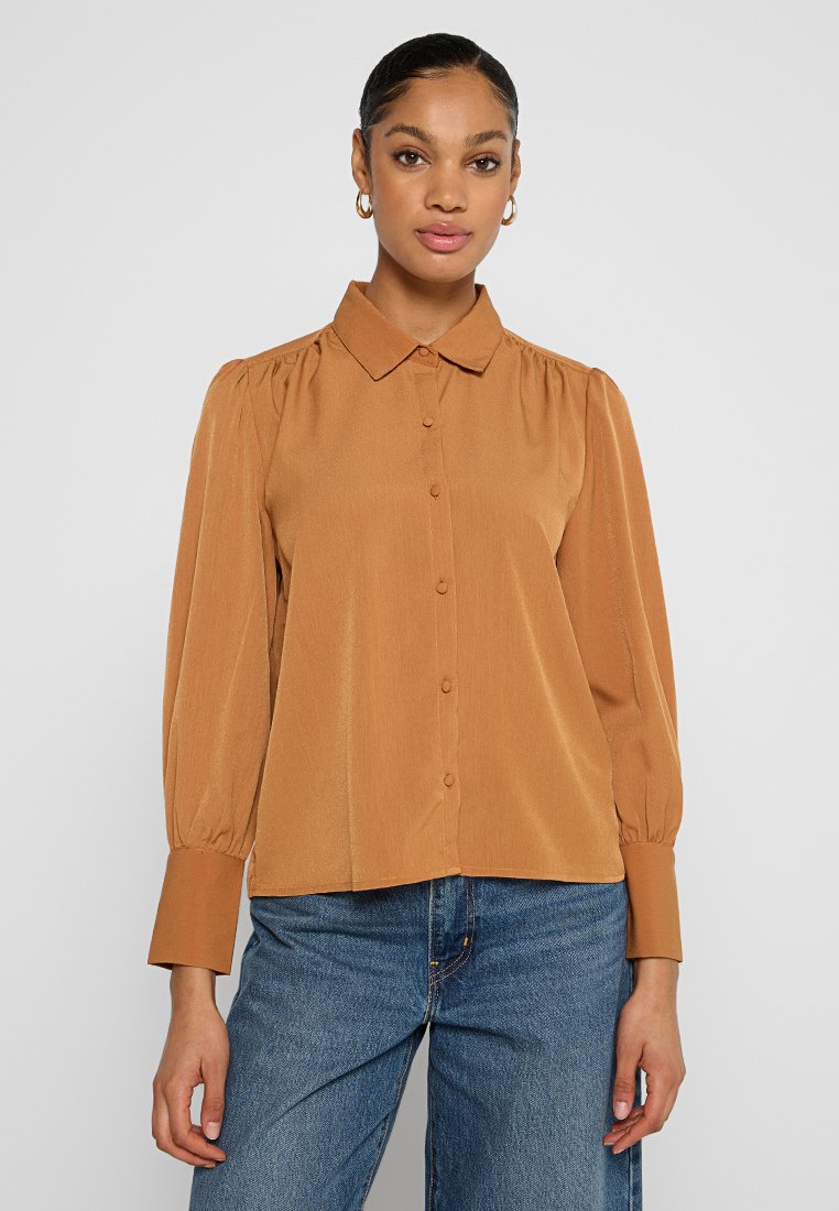 Knoop-up blouse in roestoranje, met een kraag, lange pofmouwen en een subtiele textuur. Gecombineerd met blauwe denim jeans.