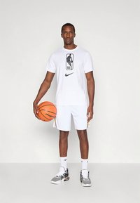 Muškarac u bijelom NBA majici i kratkim hlačama drži košarkašku loptu, stoji protiv obične svijetle pozadine, nosi bijele čarape i sive tenisice.