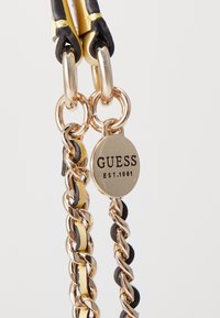 Catena in metallo tono oro con accenti neri e gialli, caratterizzata da un'etichetta circolare con la scritta "GUESS EST.1981." Finitura liscia, maglie rotonde.