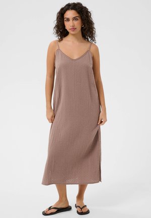 Kobieta z kręconymi włosami, ubrana w bezrękawnikową, maksi sukienkę w kolorze mauve z cienkimi ramiączkami i czarnymi klapkami, stojąca na jednolitym tle.