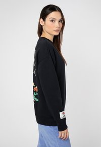 Schwarzer Pullover mit floralem Grafikdruck auf dem Rücken, gerippten Abschlüssen an den Ärmeln und am Bund. Hergestellt aus weichem Baumwollmix. Hellblaue Cordhosen.