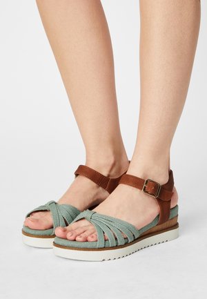 Pieds portant des sandales en tissu vert avec des brides de cheville marron et des boucles dorées, semelles blanches et un léger talon compensé sur fond blanc.
