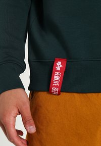Zöld pulóver bordás textúrával, a szegélyén egy "REMOTE BEFF" feliratú piros címkével, narancssárga nadrággal párosítva.