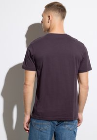 Camiseta de manga corta y cuello redondo en tejido de color púrpura oscuro, con una textura suave. Se lleva con jeans azules, mostrando un ajuste relajado.
