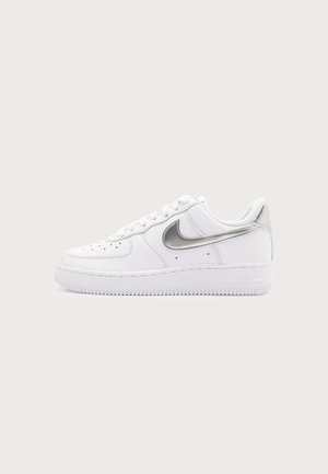 Baskets basses blanches avec le swoosh Nike argenté et l'avant perforé, présentées sur un fond gris clair uni.
