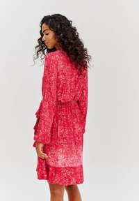 Červené květinové šaty s volným střihem, dlouhými rukávy, zavazovacími pasem a zdobeným lemováním s volánky. Materiál vypadá lehce.