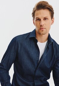 Ciemnoniebieska koszula z guzikami i kołnierzykiem, z długimi rękawami i luźnym krojem, noszona na białej koszulce.