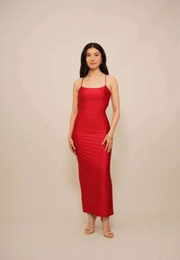 Vrouw met lang zwart haar, gekleed in een aansluitende rode mouwloze jurk met dunne bandjes en beige open teen pumps, staande tegen een effen achtergrond.