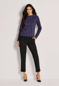 Vrouw poseert in zwarte hoge taille broek, een paars en zwart houndstooth patroon langemouwen top, zwarte puntige hakken en zilveren oorbellen.