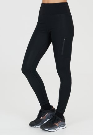 Black leggings with a high waist, smooth texture, and side zip pocket; worn with black athletic shoes featuring a rose gold accent.