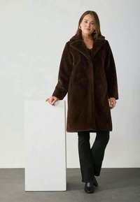Palton maro din blană artificială cu guler larg, mâneci lungi și croi drept. Purtați peste o ținută neagră, pe un fundal neutru.