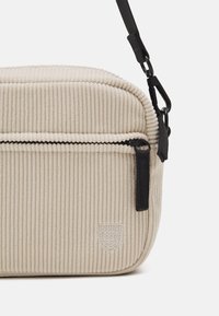 Borsa in corduroy beige con chiusura con zip e una strap nera. Caratterizzata da una superficie testurizzata e un piccolo logo impresso.