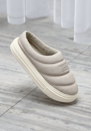 Beige slip-on sko med quiltet overdel, myk tekstur, rund form og hvit gummisåle. Har et logopatch på siden.