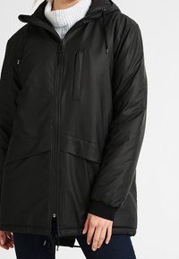 Person som bär en svart huvjacka för vintern över en vit stickad polotröja, står med avslappnade händer.