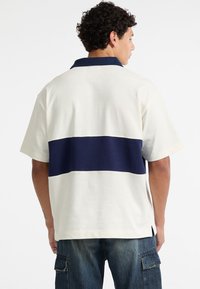 Short-sleeve shirt in cream with a horizontal navy stripe across the back. Fabric appears soft with a classic collar and side slits.