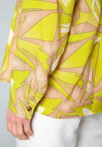 Bunt gemusterte Bluse mit einem großen grünen und beige abstrakten Muster, langen Ärmeln und einem abgerundeten Saum mit einem braunen Knopf am Ärmelbündchen.