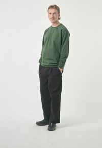 Green sweatshirt with long sleeves, black loose-fitting pants, and black shoes, standing against a plain white background.