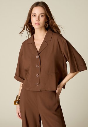 Femme portant une veste marron à manches courtes boutonnée et un pantalon assorti avec des boucles d'oreilles et bracelets dorés sur un fond uni.