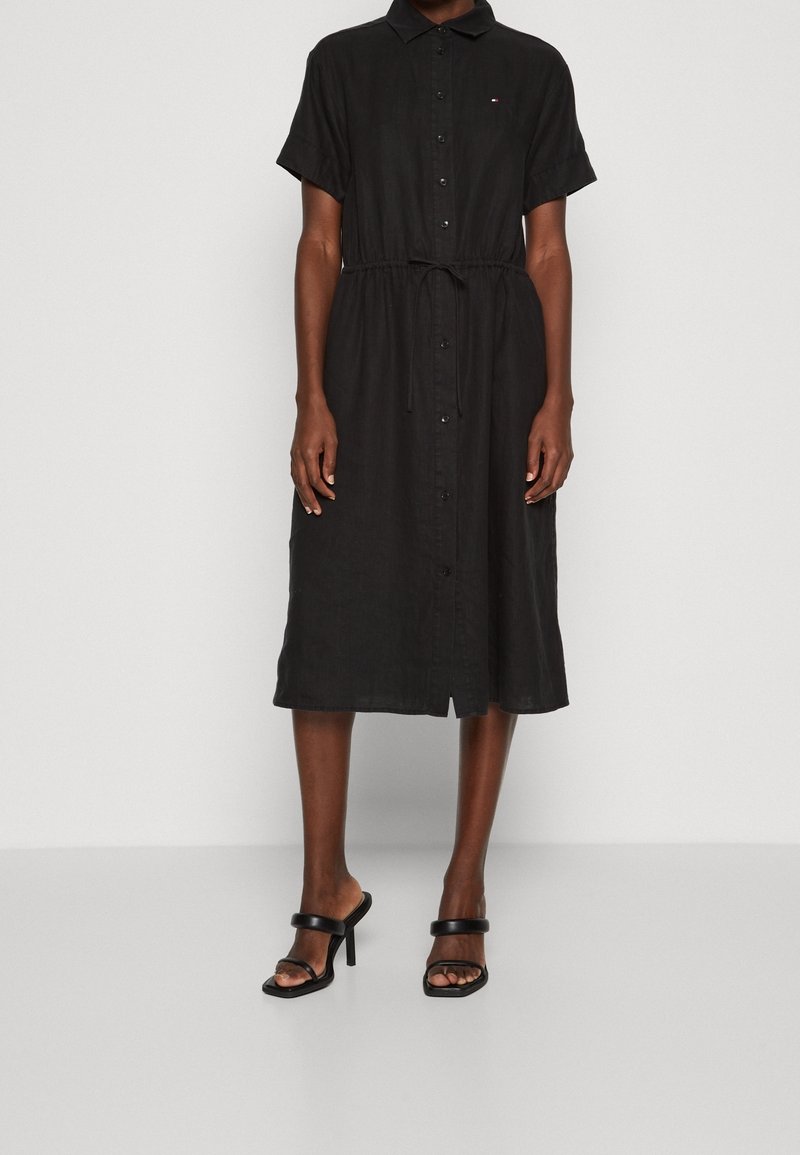 Černé košilové šaty s krátkými rukávy, páskem v pase, délkou nad kotníky a jemným logem. Vyrobené z lehké látky.