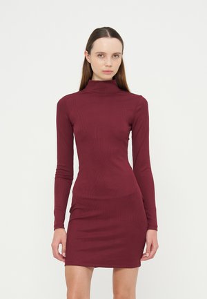 Frau steht und trägt ein figurbetontes, langärmeliges, kastanienbraunes Kleid mit hohem Ausschnitt vor einem schlichten hellen Hintergrund.