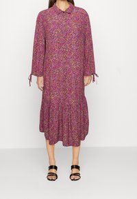 Abito floreale stampato in rosa, viola e blu. Presenta un colletto, maniche lunghe con nastri e un orlo a strati. Indossato con sandali neri con cinturini.
