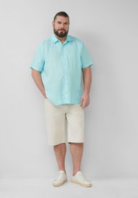 Lichtblauw linnen shirt met korte mouwen en knoopsluiting, gecombineerd met beige knielange shorts en witte sneakers op een effen achtergrond.