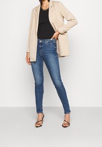 Blazer beige sobre una blusa negra, combinado con jeans ajustados azules. Los jeans tienen un ligero desgaste y detalles de costura sutiles, complementados con tacones negros de tiras.