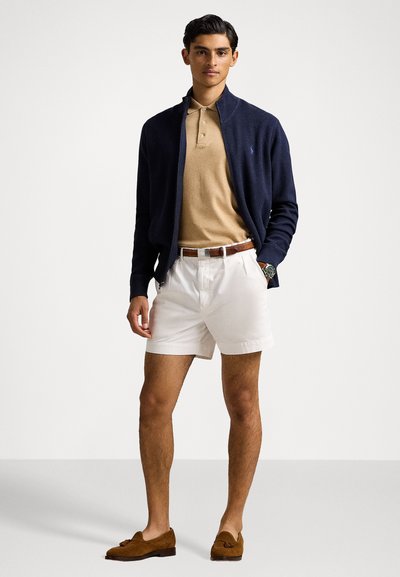 Pulover cu fermoar navy, polo bej, pantaloni scurți albi, curea maro și pantofi lejeri maro cu franjuri. Texturi fine, design ajustat și stil casual.