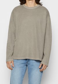 Beige ribbad långärmad tröja med rund halsringning och nedflyttade axlar, kombinerad med ljusblå jeans. Avslappnad passform och mjuk textur.